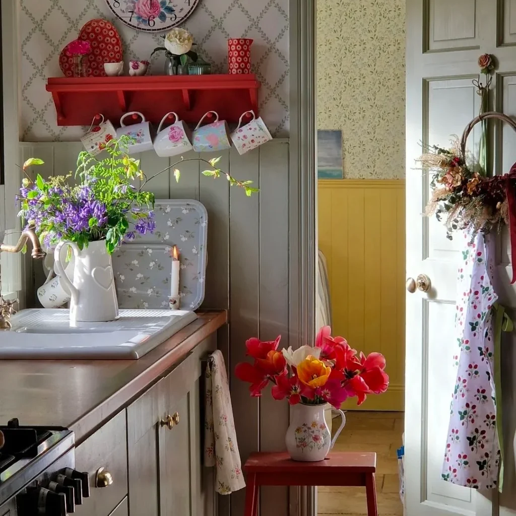 A granny chic kitchen with lots of flowers is shown. The door has flowers hanging, a vase with purple flowers on the kitchen counter, and a low wooden table with a white vase with pink flowers.