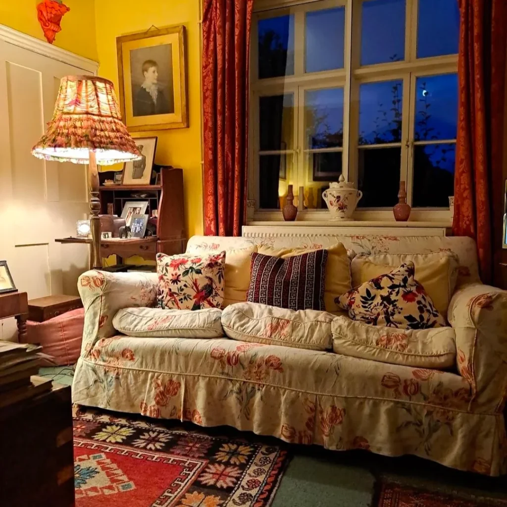 There is a very cozy, comfortable granny chic living room with maroon curtains and off white sofa covers with the same cushions. a corner, lamp and lots of vintage pieces are here.