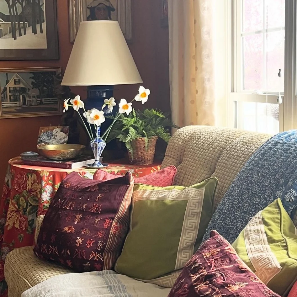 lots of cushions are placed on the sofa having a grey throw as well. a side coffee table having vase, lamp and traditional pieces is also there.