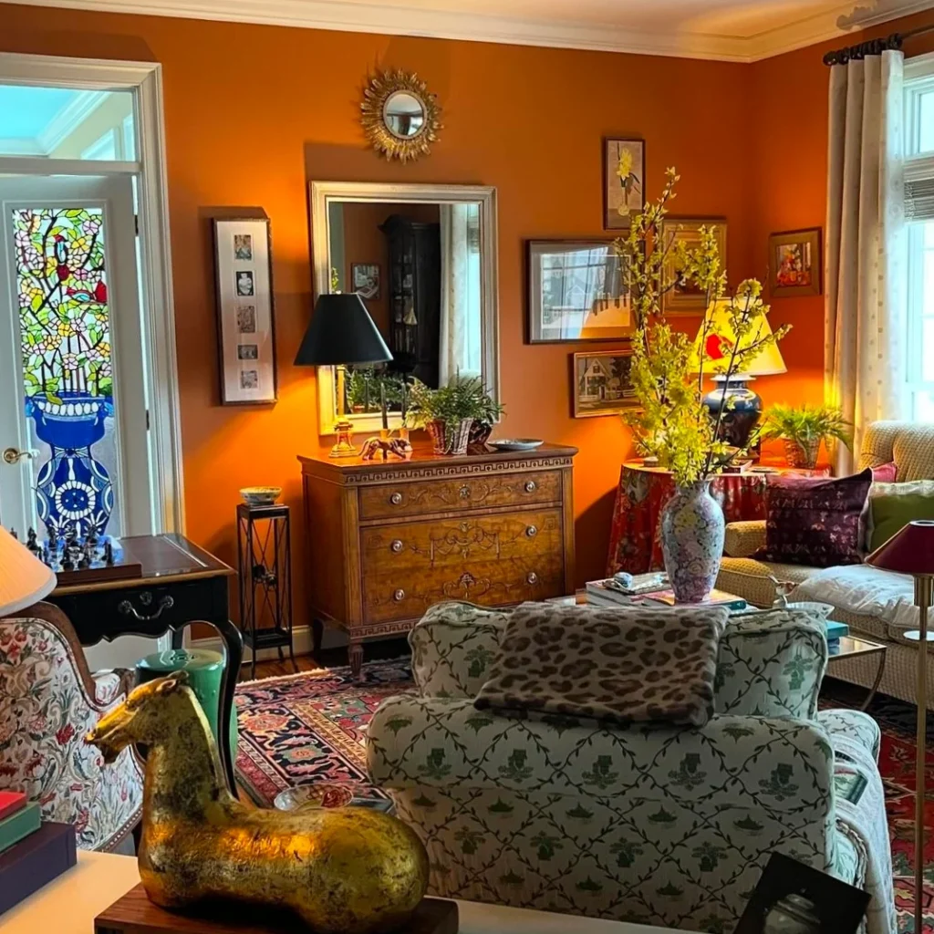 A well lighted glowing yet cozy granny chic living room with lots of wall hangings, console, wall mirror , framed pictures, a table with chess and hand painted glass door is shown.