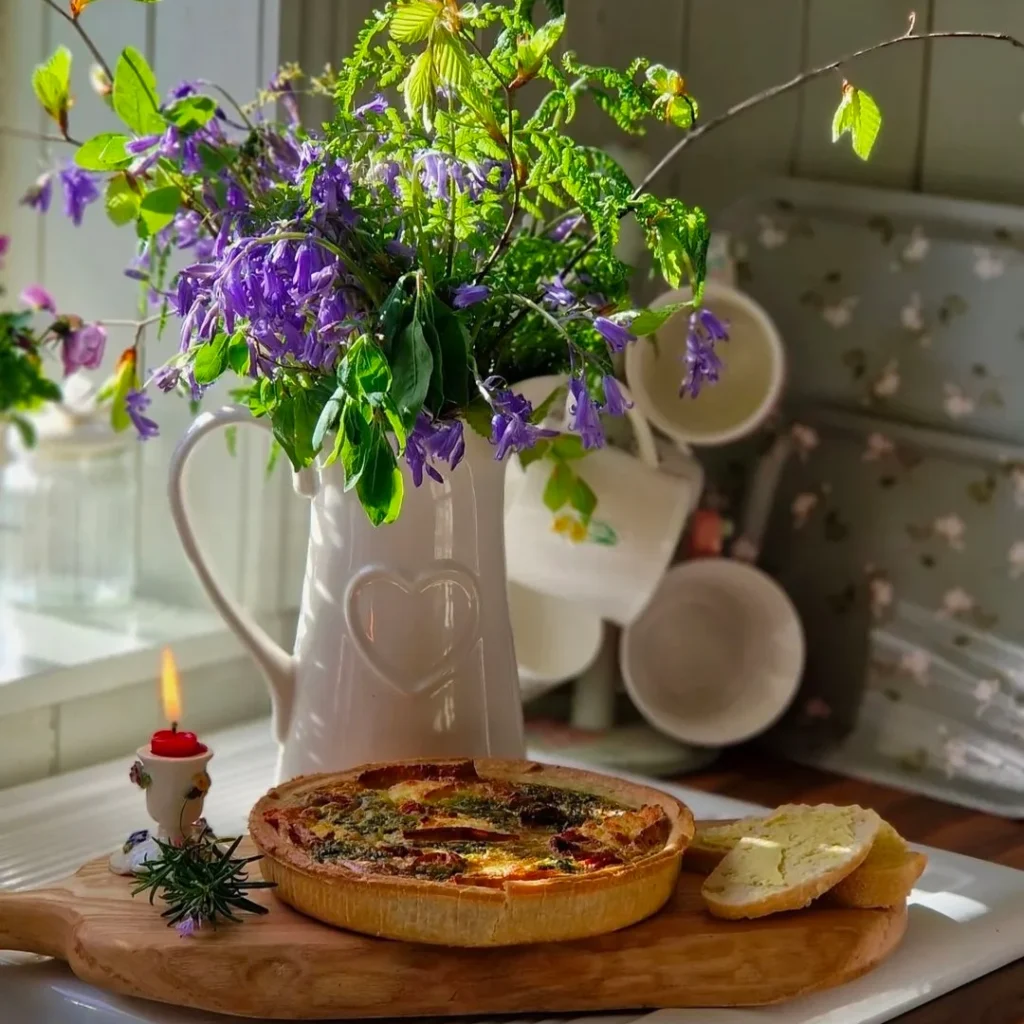 A modern styled jug shaped vase with purple and green flowers is placed on wooden chopping board. A small lighted candle bread and pizza is also placed on it.