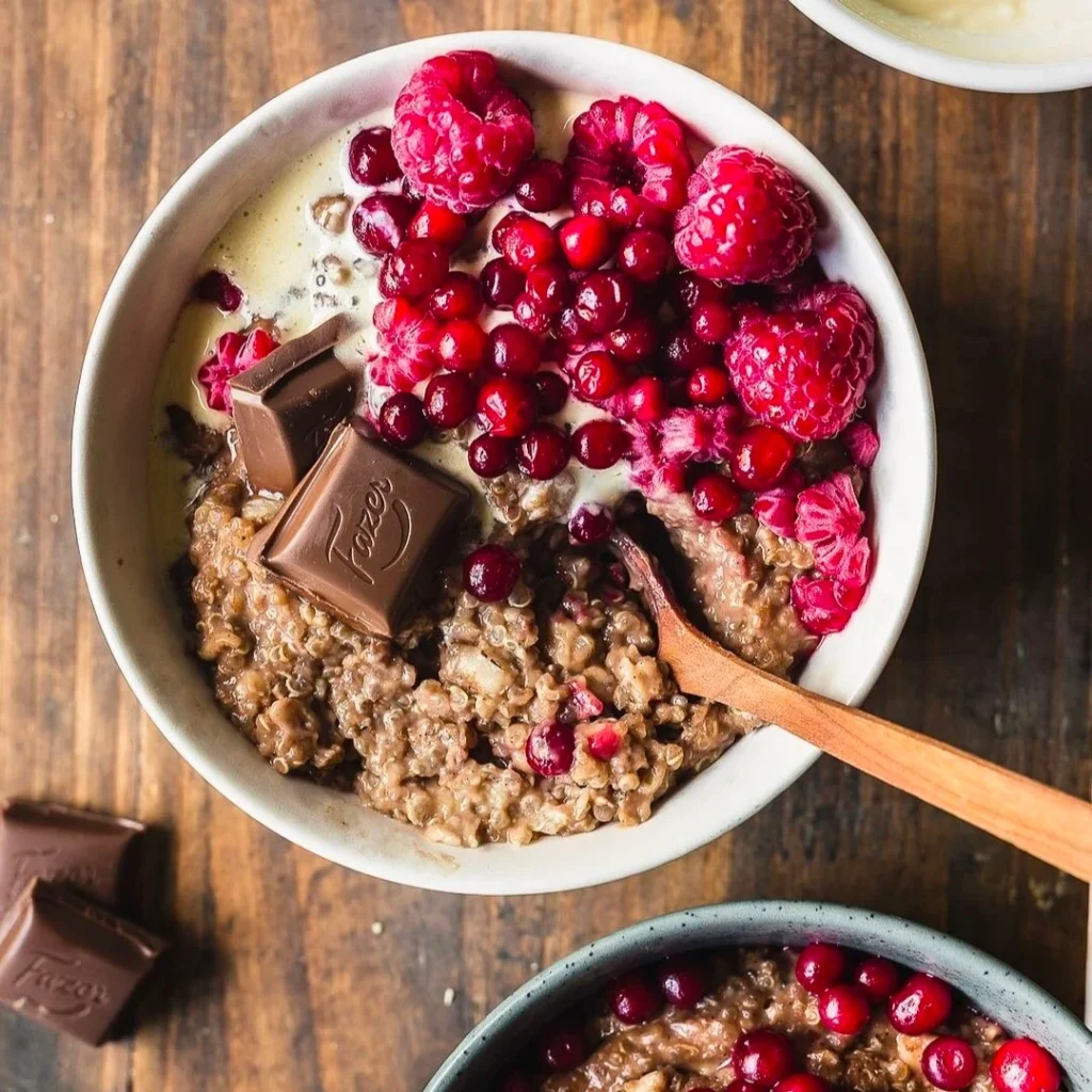 quinoa porridge in white bowl having berries, chocolate and nuts. a wooden spoon in the bowl.