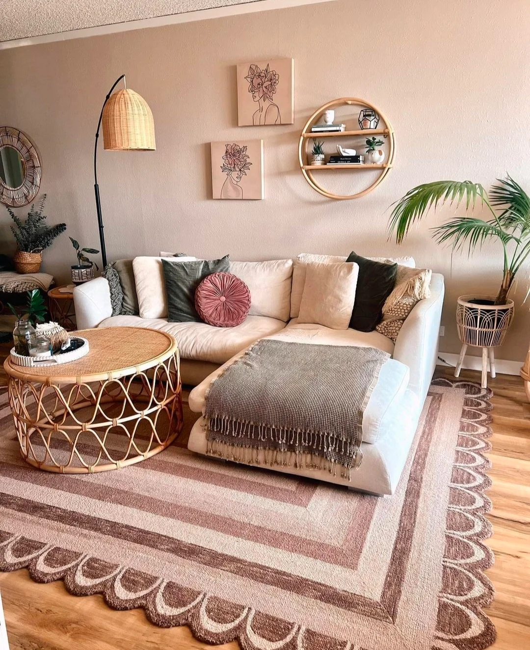 A white and brown-themed boho chic living room ideas, is decorated with a canned round table, side lamp, plant pot, L-shaped small slow-slung white sofa, and two wall hangings in a beige tone in the background.