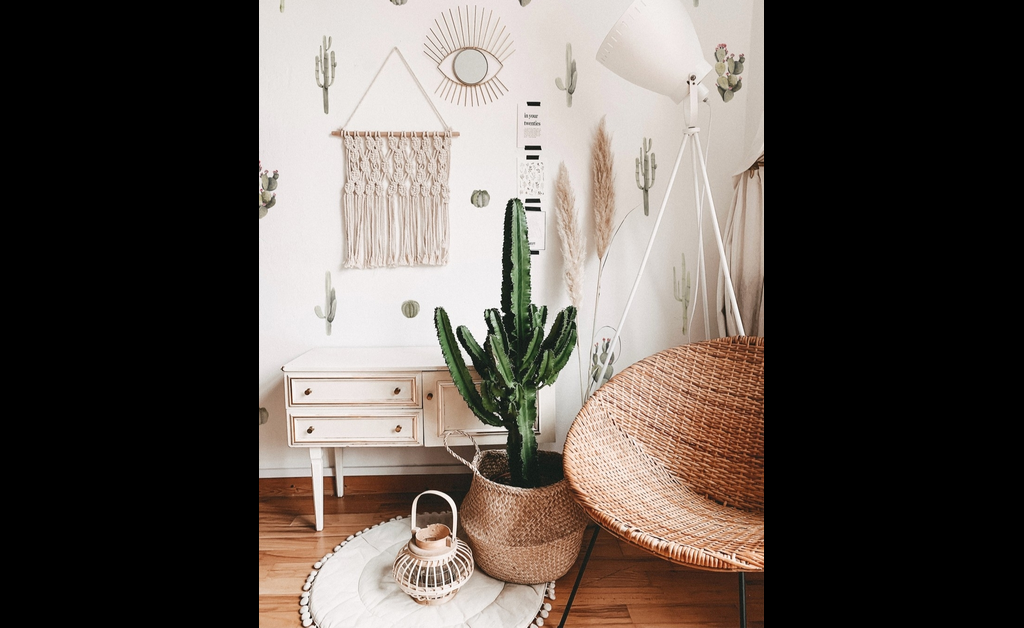 A tall saguaro cactus in woven rattan basket is placed on white round rug near a rattan egg chair.